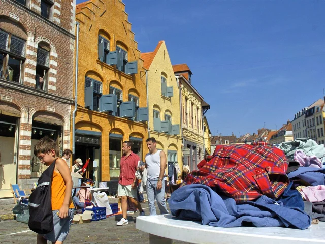Scène de braderie sous un ciel bleu à Lille, avec des passants marchant le long de bâtiments anciens en briques colorées. Au premier plan, un tas de vêtements d’occasion est posé sur une table, tandis qu’un enfant regarde le sol en portant un sac noir. En arrière-plan, plusieurs personnes déambulent entre les étals installés dans la rue pavée.