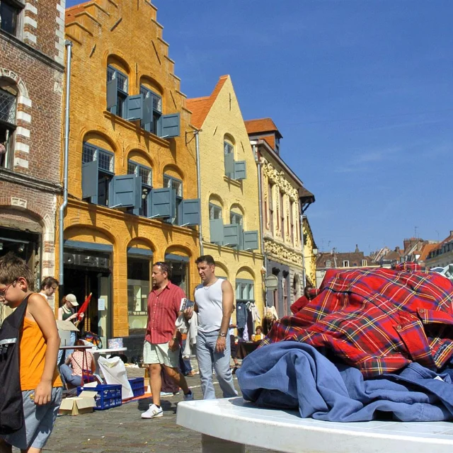 Scène de braderie sous un ciel bleu à Lille, avec des passants marchant le long de bâtiments anciens en briques colorées. Au premier plan, un tas de vêtements d’occasion est posé sur une table, tandis qu’un enfant regarde le sol en portant un sac noir. En arrière-plan, plusieurs personnes déambulent entre les étals installés dans la rue pavée.