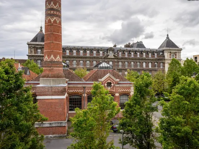 Cheminée en brique rouge ornée de motifs géométriques devant les bâtiments historiques de l'École nationale des arts et métiers de Lille, entourée d’arbres et sous un ciel nuageux.