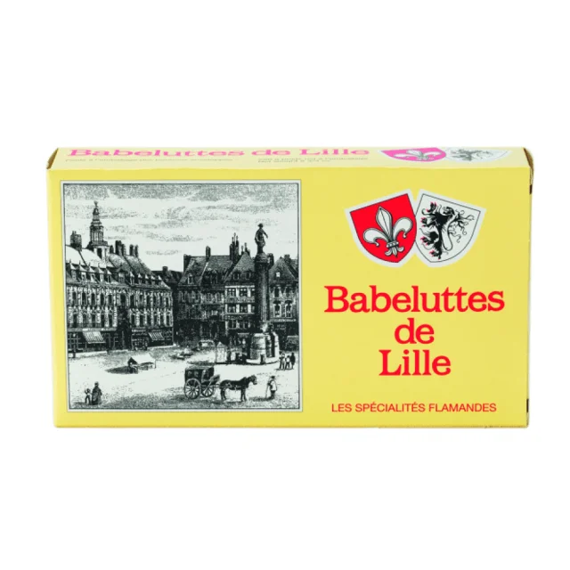 Une boîte rectangulaire jaune de Babeluttes de Lille, spécialité flamande. Sur l’emballage, une gravure en noir et blanc représente la Grand-Place de Lille avec des bâtiments anciens et une statue centrale. Deux blasons sont visibles : l’un avec une fleur de lys, l’autre avec un lion noir. Le texte rouge indique Babeluttes de Lille – Les spécialités flamandes