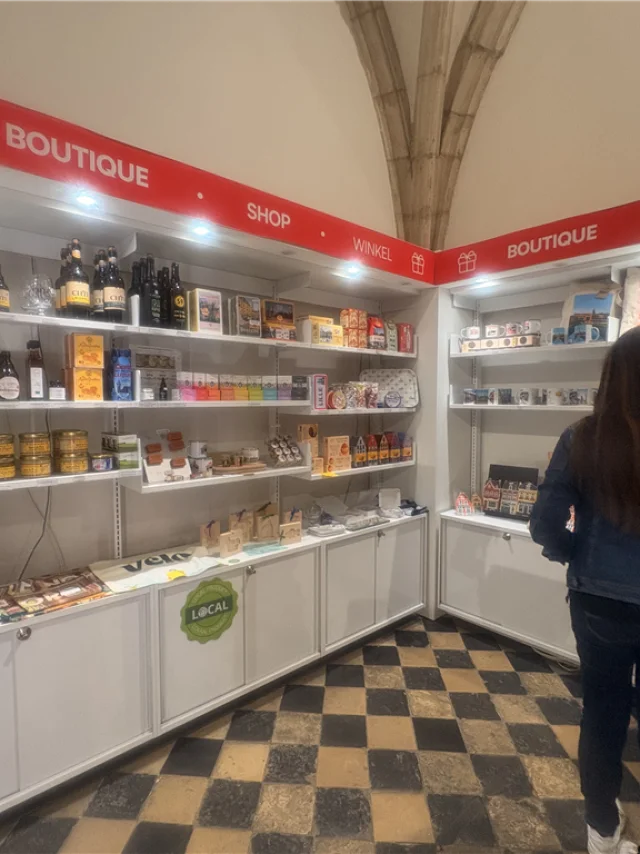 La photo montre l’intérieur d’une boutique touristique, avec des rayonnages garnis de produits locaux et souvenirs de Lille et de la Métropole : mugs, cartes postales, spécialités régionales, magnets, objets à l’effigie de la Braderie de Lille, etc. L’ambiance est lumineuse et accueillante.