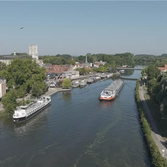 Vue aérienne du port de Wambrechies, avec ses péniches amarrées sur la Deûle, bordée de maisons et d’un château sous un ciel clair.