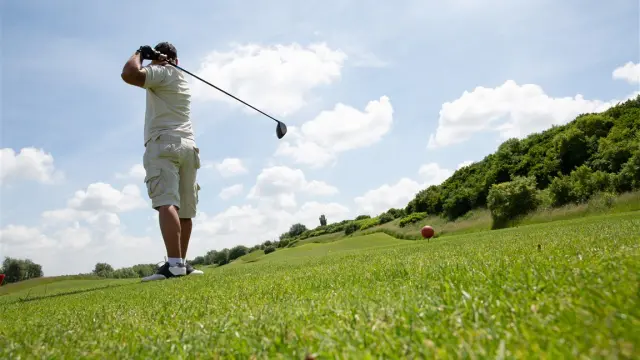 Golfeur en plein swing sur un parcours verdoyant, capturé au moment précis de l’élan. La scène s’ouvre largement sur le green vallonné, bordé de haies et de bosquets, sous un ciel lumineux ponctué de nuages, évoquant une pratique sportive au grand air, calme et maîtrisée.