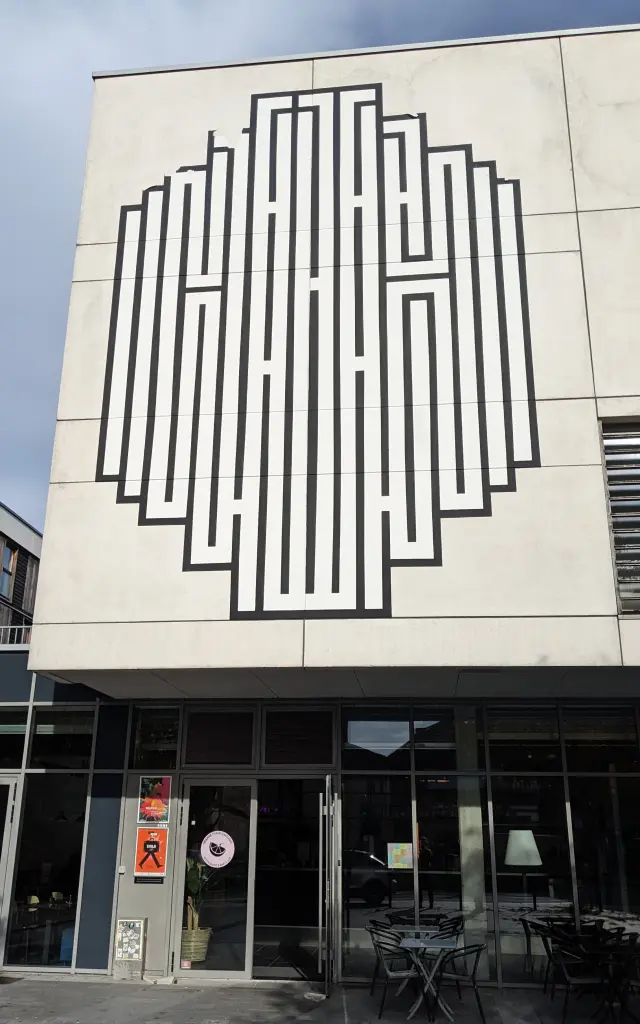 Fresque géométrique en noir et blanc de l’artiste Atlas sur la façade d’un bâtiment, composée de lignes verticales formant un motif labyrinthique.