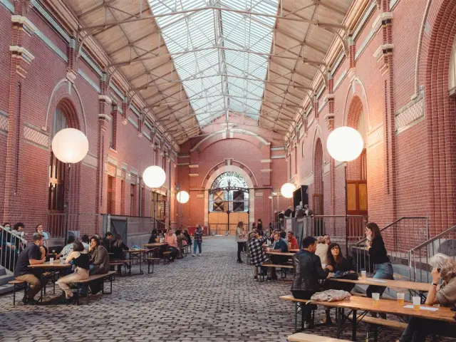 La Rue couverte de la Condition Publique à Roubaix, animée par des visiteurs discutant autour de grandes tables sous la verrière.